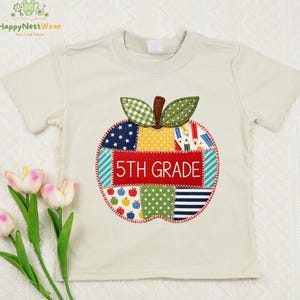 May include: A cream-colored short-sleeve t-shirt featuring a large, central apple-shaped applique made of various patterned fabric patches. The center red patch displays the white embroidered text 5TH GRADE, surrounded by colorful designs.