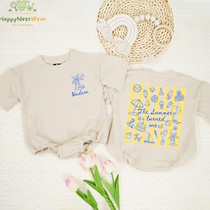 May include: Two beige baby rompers. One has a blue palm tree and the name "Hudson" printed on the front. The other has a yellow and blue summer-themed graphic with the text "The Summer i turned ones".