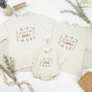 May include: Three off-white t-shirts and a onesie with colorful text and graphics. The shirts say "It's fun to be dada," "It's fun to be mama," and the onesie says "It's fun to be one." The shirts and onesie have birthday-themed designs.