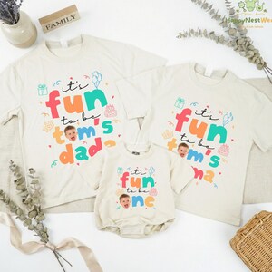 May include: Off-white family matching t-shirts and a onesie with the text "It's fun to be Tom's Dad", "It's fun to be Tom's Mom", and "It's fun to be one". The shirts have colorful graphics and a photo of a baby.