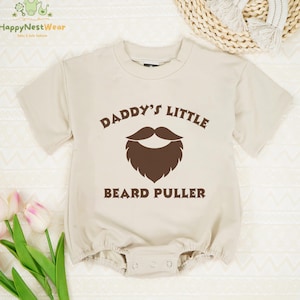 May include: Beige baby onesie with the text "DADDY'S LITTLE BEARD PULLER" and a brown beard graphic. The onesie has short sleeves and a round neckline. The onesie has snap closures at the bottom.