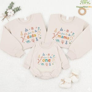 May include: Three beige sweatshirts and a romper with colorful embroidered text and graphics. The sweatshirts read "It's fun to be dada" and "It's fun to be mama". The romper says "It's fun to be one". The image also includes a pair of white baby booties.