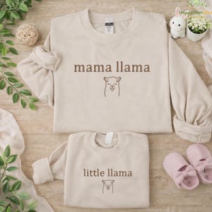 May include: Two beige sweatshirts, one adult and one child size, with the text "mama llama" and "little llama" in brown. Each sweatshirt features a llama graphic. Pink baby shoes are next to the child's sweatshirt.