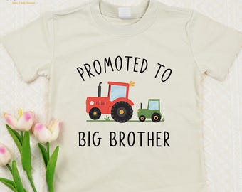May include: A cream-colored short-sleeve t-shirt featuring a graphic of a large red tractor and a smaller green tractor. The text "PROMOTED TO" is printed in black above the tractors, and the text "BIG BROTHER" is printed in black below them.