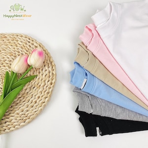 May include: A stack of colorful crew neck sweatshirts in white, pink, tan, light blue, gray, and black. The sweatshirts are neatly arranged, showcasing their different hues. The logo "HappyNestWear" is visible in the top left corner.