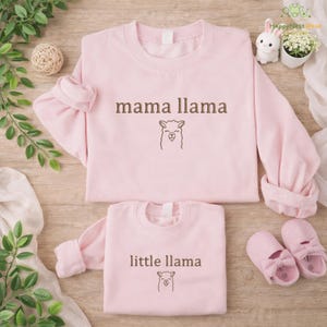 May include: Two pale pink sweatshirts, one adult and one child size, featuring the text "mama llama" and "little llama" in brown, each with a llama graphic. The image also includes pink baby shoes and decorative items.