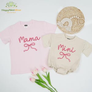 May include: A light pink t-shirt with the word "Mama" in pink script and a pink bow graphic. Beside it is a beige baby romper with the word "Mini" in pink script and a pink bow graphic. A woven mat and a rainbow toy are in the background.