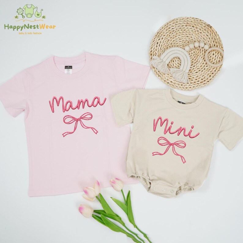 May include: A light pink t-shirt with the word "Mama" in pink script and a pink bow graphic. Beside it is a beige baby romper with the word "Mini" in pink script and a pink bow graphic. A woven mat and a rainbow toy are in the background.