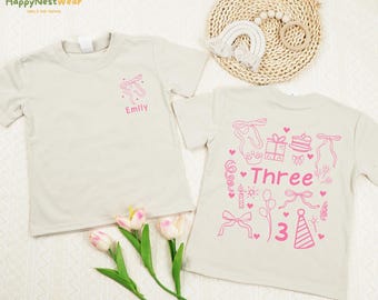May include: Two beige children's t-shirts. One shirt has a pink ballet shoe design and the name "Emily". The other shirt has a pink "Three" birthday design with balloons, a cake, and a party hat.