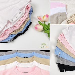 Op de afbeelding: Een collectie babykleding in diverse kleuren, waaronder wit, roze, beige, blauw, grijs en zwart. De afbeelding toont verschillende rompertjes met drukknoopsluitingen en crewneck sweatshirts. Het HappyNestWear-logo is zichtbaar.
