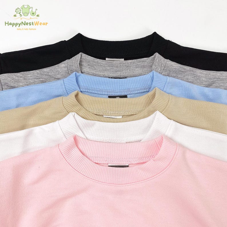 May include: A close-up shot of several crew neck sweatshirts in various colors. The colors include black, gray, light blue, tan, white, and pink. The sweatshirts have ribbed collars and cuffs. The brand name "HappyNestWear" is visible in the top left corner.