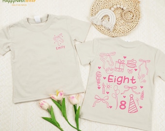 May include: Two beige t-shirts with pink designs. One shirt has the name "Emily" and a bow design. The other shirt says "Eight" with birthday-themed graphics. The shirts are on a white surface with flowers and a woven mat.