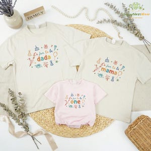 May include: Family matching t-shirts and a baby onesie. The t-shirts are off-white with the text "It's fun to be dada" and "It's fun to be mama" in colorful embroidery. The baby onesie is pink and says "It's fun to be one".
