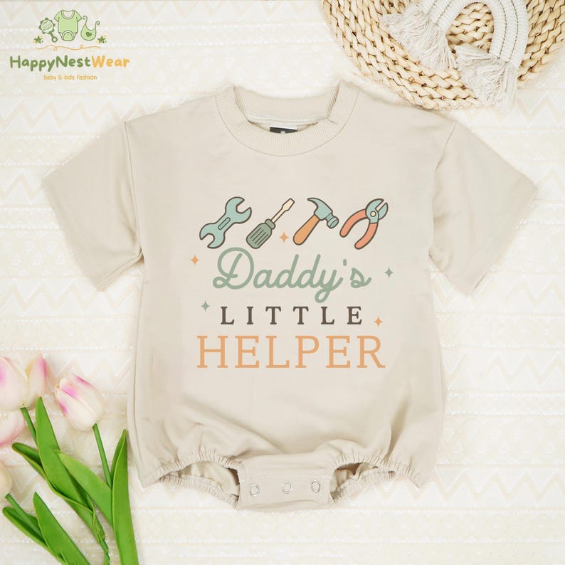 May include: Beige baby romper with the text "Daddy's Little Helper" and tool graphics. The short-sleeved one-piece features a crew neck and snap closure. The romper is made of a soft, comfortable material.