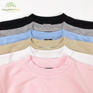Puede incluir: Primer plano de varias sudaderas de cuello redondo en varios colores. Los colores incluyen negro, gris, azul claro, beige, blanco y rosa. Las sudaderas tienen cuellos y pu&ntilde;os acanalados. La marca "HappyNestWear" es visible.