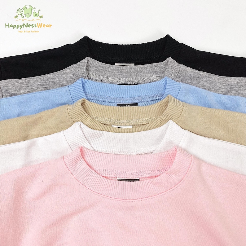 Puede incluir: Primer plano de varias sudaderas de cuello redondo en varios colores. Los colores incluyen negro, gris, azul claro, beige, blanco y rosa. Las sudaderas tienen cuellos y pu&ntilde;os acanalados. La marca "HappyNestWear" es visible.
