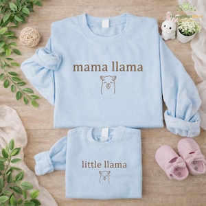 May include: Two pale blue sweatshirts, one reading "mama llama" and the other "little llama" in brown text, each with a llama illustration. The sweatshirts are on a wooden surface, with a decorative ball, a bunny figurine, and pink baby shoes.