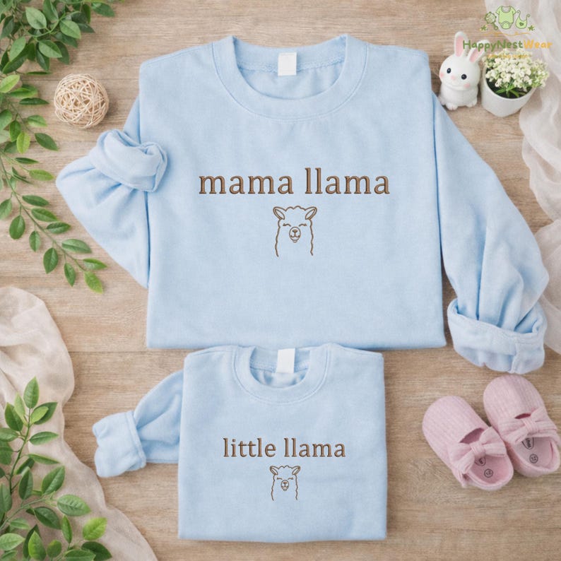 May include: Two pale blue sweatshirts, one reading "mama llama" and the other "little llama" in brown text, each with a llama illustration. The sweatshirts are on a wooden surface, with a decorative ball, a bunny figurine, and pink baby shoes.