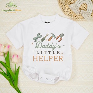 May include: White baby onesie with short sleeves and a round neckline. The onesie features a graphic of tools and the text "Daddy's Little Helper" in green and brown. The garment has snap closures at the bottom.