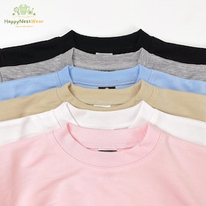 Op de afbeelding: Een close-up van een stapel crewneck sweatshirts in verschillende kleuren: zwart, grijs, lichtblauw, beige, wit en roze. De sweatshirts zijn netjes gerangschikt en tonen hun geribbelde kragen en zachte stof. Het HappyNestWear-logo is zichtbaar.
