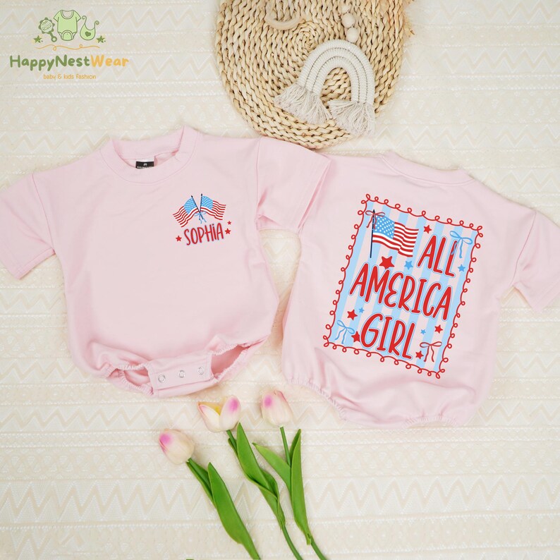 Puede incluir: Dos peleles rosas para beb&eacute;s. Uno presenta el nombre "SOPHIA" con banderas americanas. El otro dice "ALL AMERICA GIRL" con estrellas y una bandera americana. Ambos tienen mangas cortas y cierre de broche.