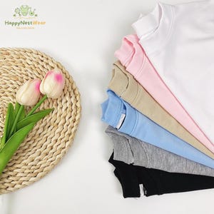 Puede incluir: Una pila de sudaderas de cuello redondo de colores: blanco, rosa, beige, azul claro, gris y negro. Las sudaderas est&aacute;n cuidadosamente dispuestas, mostrando sus diferentes tonos. El logotipo "HappyNestWear" es visible en la esquina superior izquierda.