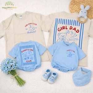 May include: A collection of baby clothing and accessories in soft pastel colors. Includes beige t-shirts with "GIRL DAD CLUB" and "DADDY'S GIRL" graphics, blue rompers, matching shoes, and a bib. A wooden ring toy and a bouquet of blue flowers are also present.