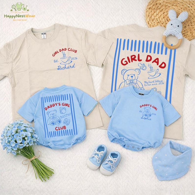 May include: A collection of baby clothing and accessories in soft pastel colors. Includes beige t-shirts with "GIRL DAD CLUB" and "DADDY'S GIRL" graphics, blue rompers, matching shoes, and a bib. A wooden ring toy and a bouquet of blue flowers are also present.
