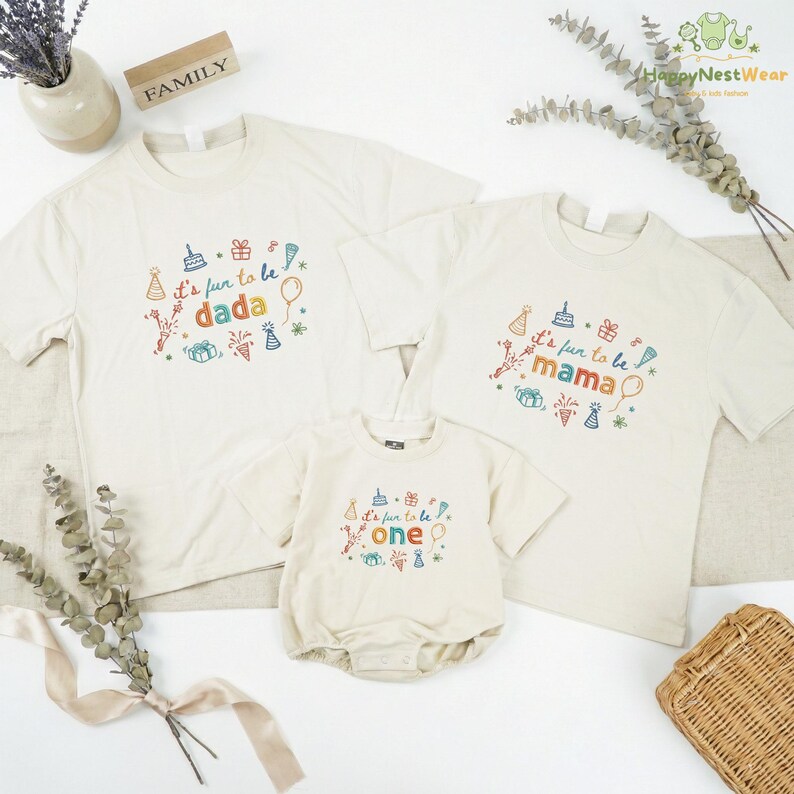 May include: Three off-white t-shirts and a onesie with colorful text and graphics. The shirts say "It's fun to be dada," "It's fun to be mama," and the onesie says "It's fun to be one." The shirts and onesie have birthday-themed designs.