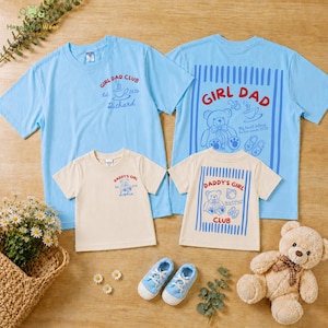 May include: Four light blue and cream-colored t-shirts with text and graphics. The shirts say "GIRL DAD CLUB" and "DADDY'S GIRL CLUB". There are also baby shoes and a teddy bear. The shirts are for a family.
