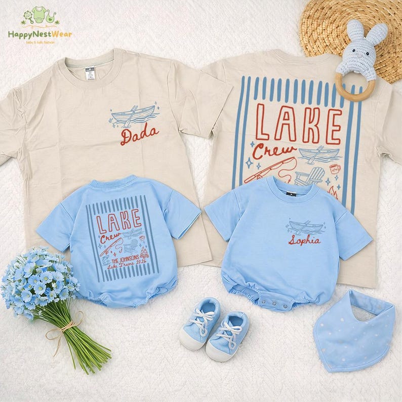 May include: A collection of light blue and beige baby clothing and accessories. Includes t-shirts, a romper, shoes, and a bib. The shirts have lake-themed graphics and text, including "Dada," "Lake Crew," and a name. A blue floral bouquet and a bunny-shaped teether are also present.