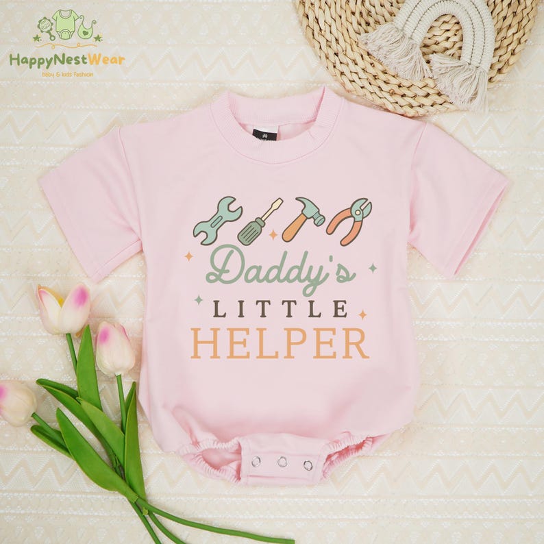 May include: A light pink baby bodysuit with short sleeves. The front features a graphic of tools and the text "Daddy's Little Helper" in green and orange. The bodysuit has snap closures at the bottom.