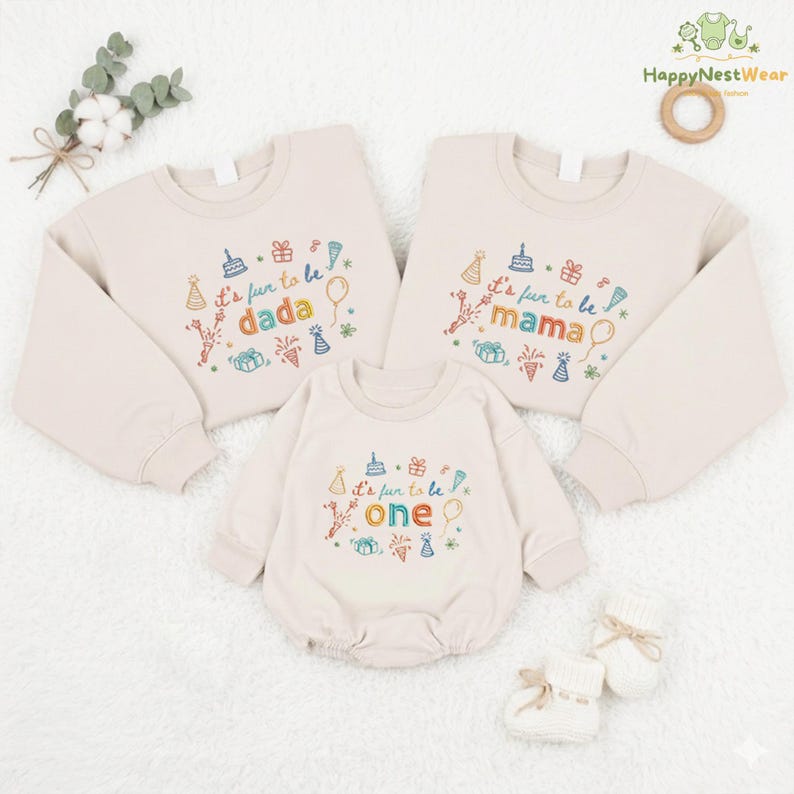May include: Three beige sweatshirts and a romper with colorful embroidered text and graphics. The sweatshirts read "It's fun to be dada" and "It's fun to be mama". The romper says "It's fun to be one". The image also includes a pair of white baby booties.