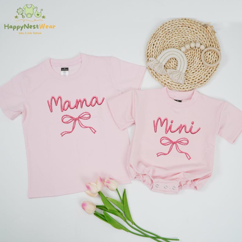 May include: Pink t-shirt with the word "Mama" embroidered in pink script and a pink bow. A matching pink onesie with "Mini" embroidered in pink script and a pink bow. A set of matching outfits.