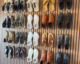 May include: A collection of various footwear arranged in rows on a slatted wall, including athletic sneakers, leather dress shoes, sandals, and boots in diverse colors such as white, black, brown, and grey, displayed in pairs.