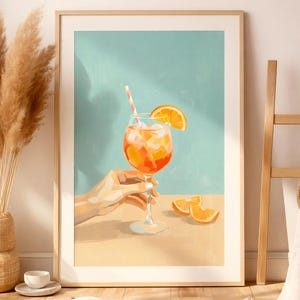 May include: A framed art print depicts a cocktail in a stemmed glass with an orange slice and a striped straw. The drink is held by a hand. The background is a light blue and tan color.