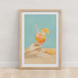 May include: A framed art print featuring a refreshing cocktail. The drink, in a stemmed glass, is orange with ice and a straw. An orange slice adorns the rim. A hand holds the glass. Slices of orange are on the table. The background is a light blue.