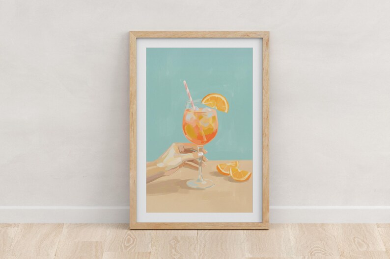 May include: A framed art print featuring a refreshing cocktail. The drink, in a stemmed glass, is orange with ice and a straw. An orange slice adorns the rim. A hand holds the glass. Slices of orange are on the table. The background is a light blue.