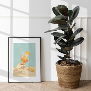 May include: A framed art print depicts a refreshing cocktail with an orange slice and a striped straw. The drink is held by a hand. The print is set against a light blue background. A large potted plant with dark green leaves sits to the right.