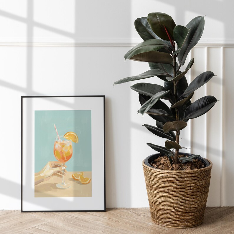 May include: A framed art print depicts a refreshing cocktail with an orange slice and a striped straw. The drink is held by a hand. The print is set against a light blue background. A large potted plant with dark green leaves sits to the right.