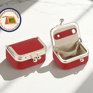 Op de afbeelding: Twee rode, rechthoekige munttasjes met zilverkleurige frames. Eén is gesloten, de andere open en onthult een beige interieur. De tasjes hebben een getextureerd oppervlak en een sluiting met kliksluiting. Het logo "Dainty Pouch & Charms" staat linksboven.