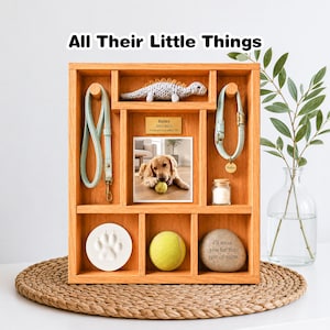 May include: A wooden display box with various compartments, containing a photo of a golden retriever, a paw print, a tennis ball, and a leash. The text "All Their Little Things" is at the top. A small dinosaur toy and a collar are also displayed.