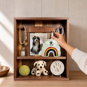 May include: A wooden shadow box memorial display featuring a dog collar, a photo of a dog, a ceramic urn with a rainbow and paw print, a tennis ball, a plush dog toy, and a clay paw print impression. The plaque reads Sammy 2009-2026.