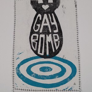 Peut inclure: Impression rectangulaire blanche avec une forme de bombe noire et les mots "GAY BOMB" en blanc. Une forme de cœur est en haut de la bombe. En dessous, des cercles concentriques bleus. L'impression a une bordure cousue.