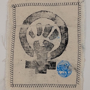 May include: A square, off-white fabric patch featuring a black, stylized image of a raised fist within a circle, above a gender symbol. It has a black stitched border and a small blue circular stamp.