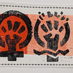 May include: A rectangular fabric patch with two black and red graphic symbols. The symbols are printed on a white and orange background. The patch is bordered with black stitching.