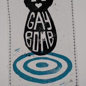 Peut inclure: Textile rectangulaire blanc avec un motif de bombe noire et les mots "GAY BOMB" en blanc. Une forme de cœur est au-dessus du texte. En dessous de la bombe, un motif de cible bleu. Les bords sont cousus avec du fil noir.