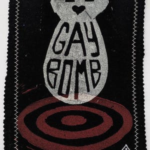 Pode incluir: Remendo de tecido preto com borda costurada branca. O remendo apresenta um design de bomba estilizado em prata com as palavras "GAY BOMB" no centro. Um design de alvo vermelho está abaixo da bomba.