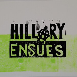 Könnte beinhalten: Ein Druck mit den Worten "HILLARY ENSUES" in schwarzen Blockbuchstaben, mit einem Anarchie-Symbol über dem "RY". Der Text befindet sich auf weißem Hintergrund mit einem grünen horizontalen Streifen unten.