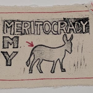 Puede incluir: Un parche rectangular con la palabra "MERITOCRACY" impresa en letras negras en bloque sobre una ilustración de un burro negro. Las letras "M" e "Y" están impresas debajo de la palabra "MERITOCRACY". Una flecha roja apunta al burro. El parche está bordeado con costuras rojas.